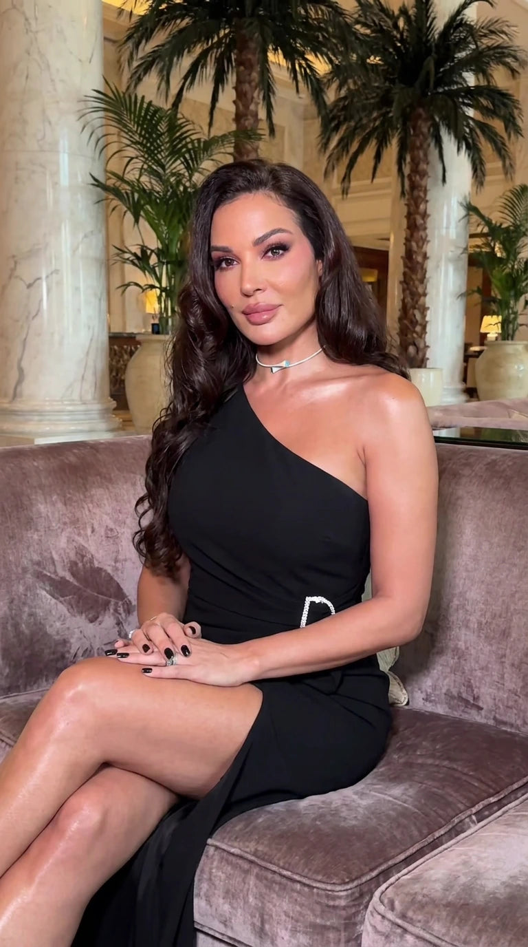 Model seated wearing a black one-shoulder evening maxi dress with waist brooch and leg slit