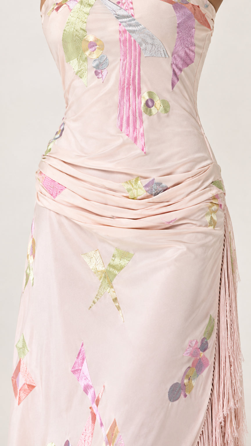 Close-up detail of blush pink dress showing gathered fabric, abstract motif, and fringe trim