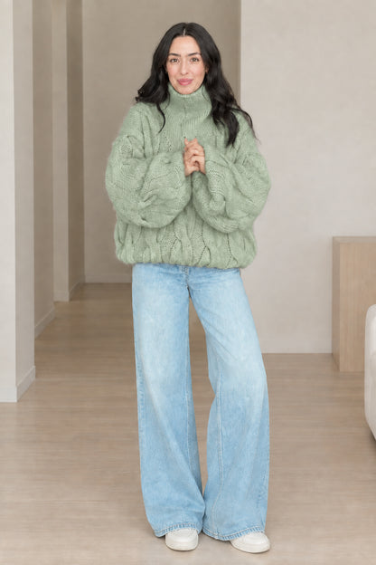 Front view of model wearing sage green chunky cable knit turtleneck sweater with wide-leg jeans